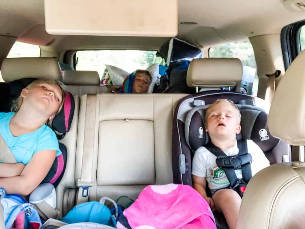 A picture taken from the front seat of a suburban backwards. 3 kids are fast asleep with their mouths hanging open. 