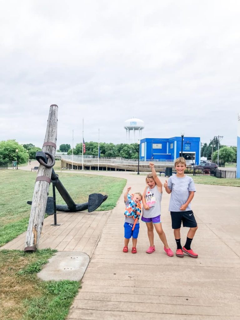 3 kids standing near a large ship anchor. In the background there is a white water tower that says Alpena on it. Blue Building in the background that is the NOAA headquarters. 