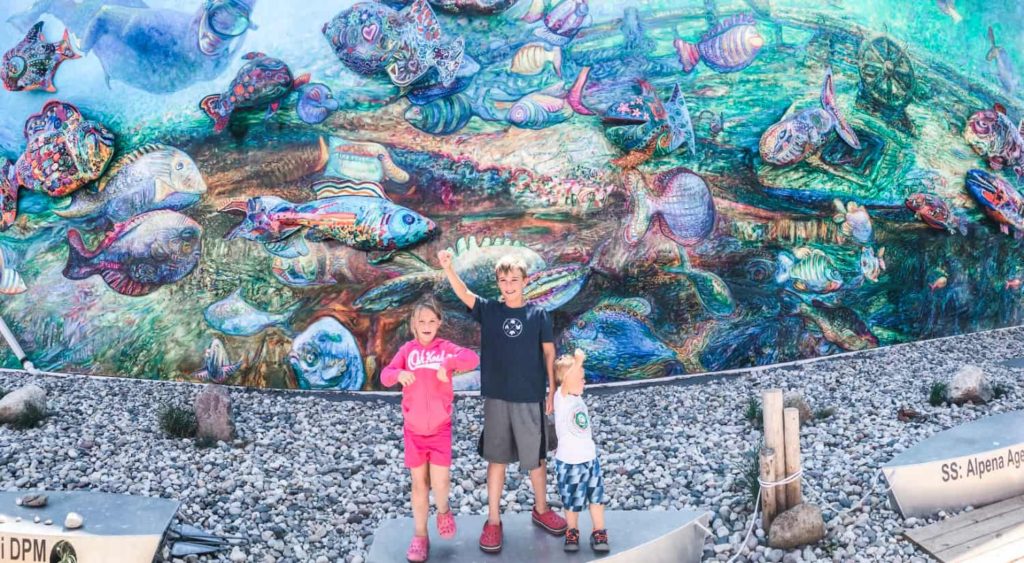 Large mural painted on the side of a building. The theme of the building is "under the sea" three kids standing in front of the wall. 