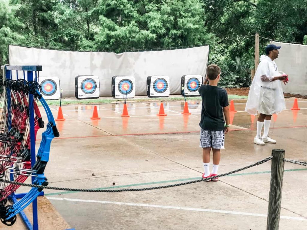 Rack of bows off to the side, archery targets lined up in the distance, instructor wearing a poncho giving directions while young boy stands and listens. 