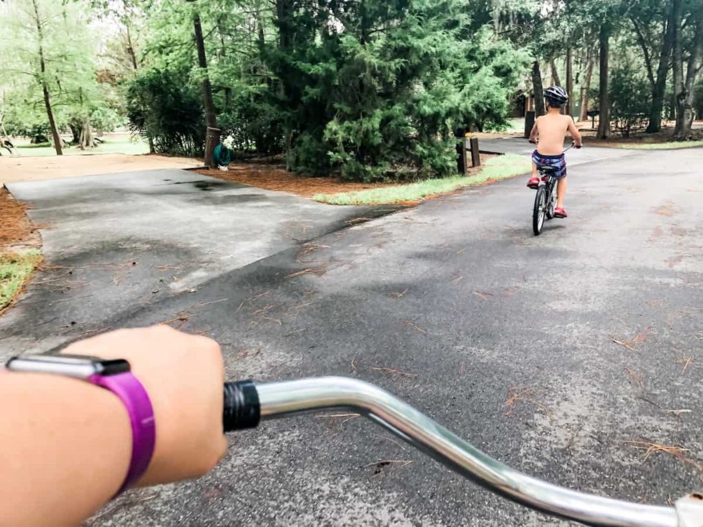 woman's hand and handlebars of her bike, as the perspective of the picture is her watching a young boy ride his bike through a paved path through the woods. 
