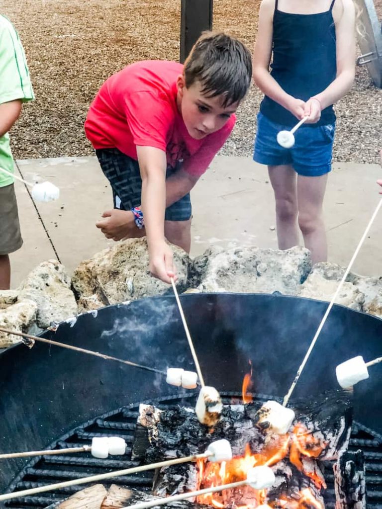 Boy roasting a marshmallow. Several other marshmallows being held by other people over the fire as well. 