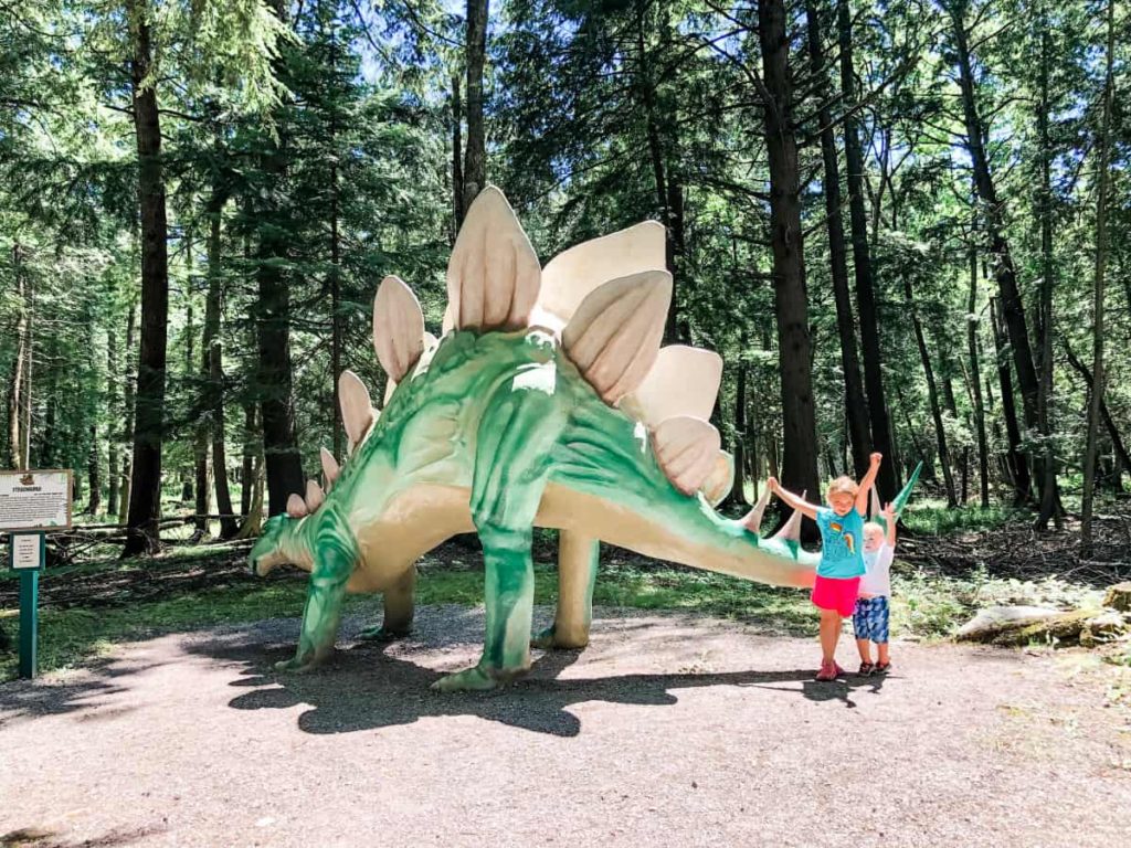 Boy and girl standing next to a giant stegosaurus statue. Statue is about 12-15 feet tall at the top of its back. 