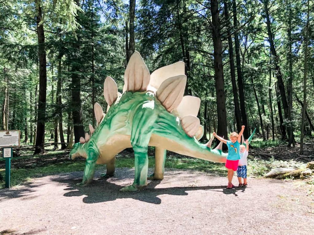 Boy and girl standing next to a giant stegosaurus statue. Statue is about 12-15 feet tall at the top of its back.