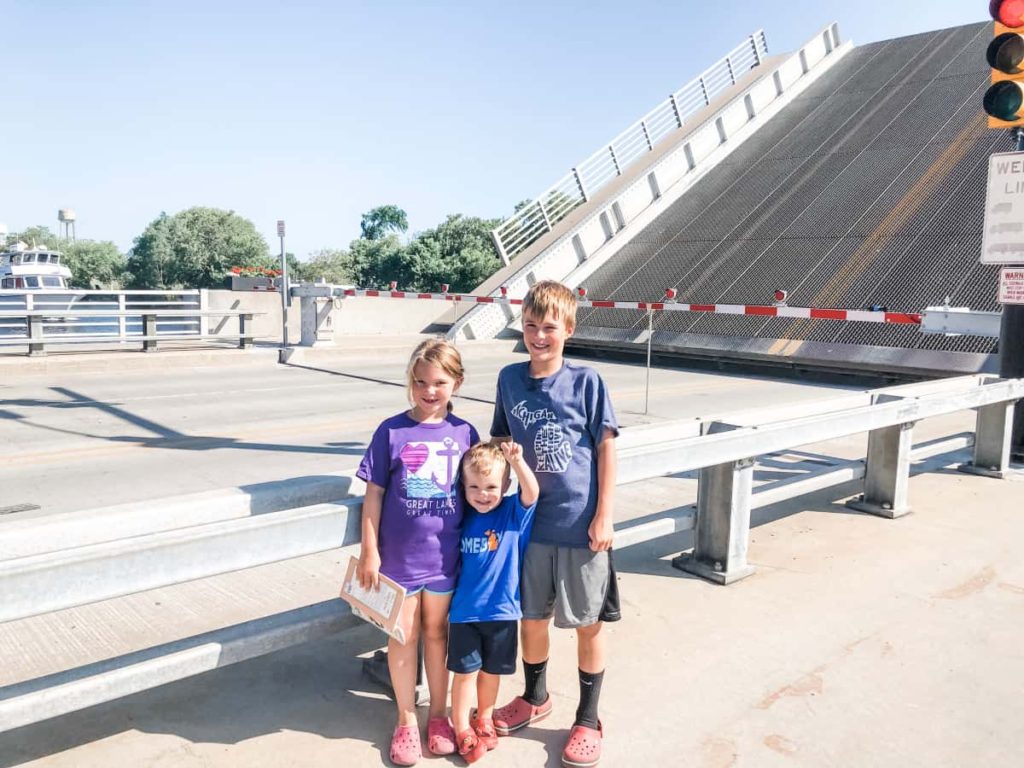 3 kids standing in front of a draw bridge. The bridge is at about a 45 degree angle. Rail Road crossing type road blocks are down to tell cars to stop. 