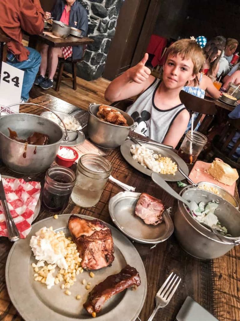 Boy giving approving thumbs up, a feast of food including ribs, chicken, mashed potatoes, corn, corn bread, all served in pewter buckets. Drinks served in mason jars. 