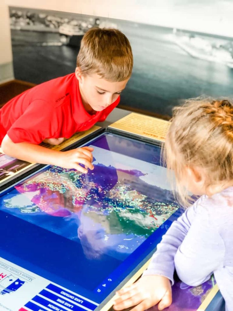 Boy and Girl looking at a flat computer screen that shows the global position of all the ships currently on the sea. 