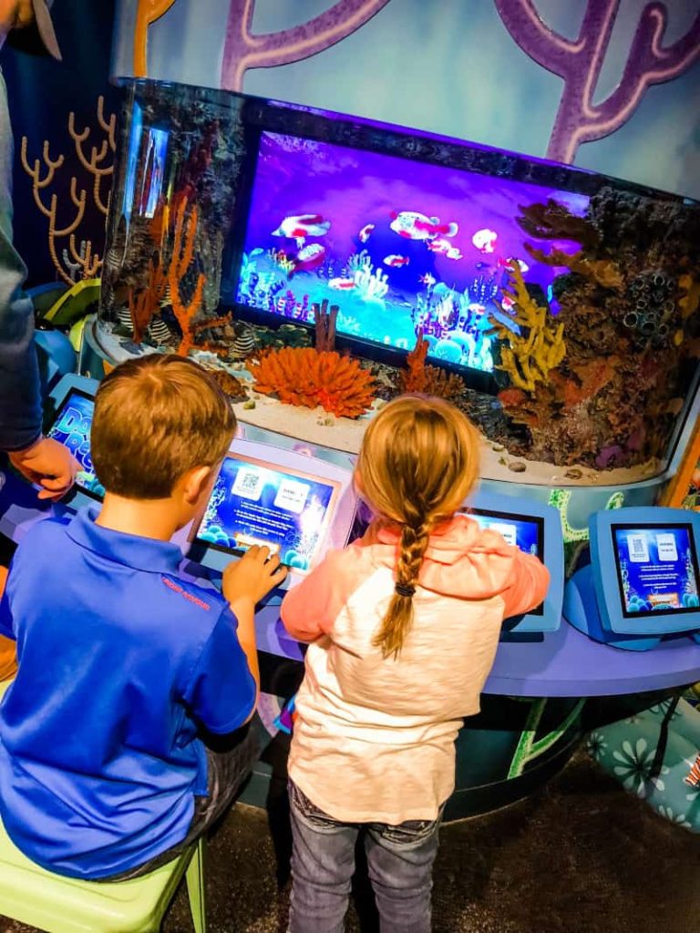 2 children playing on tablets creating digital fish that are being displayed on a large screen