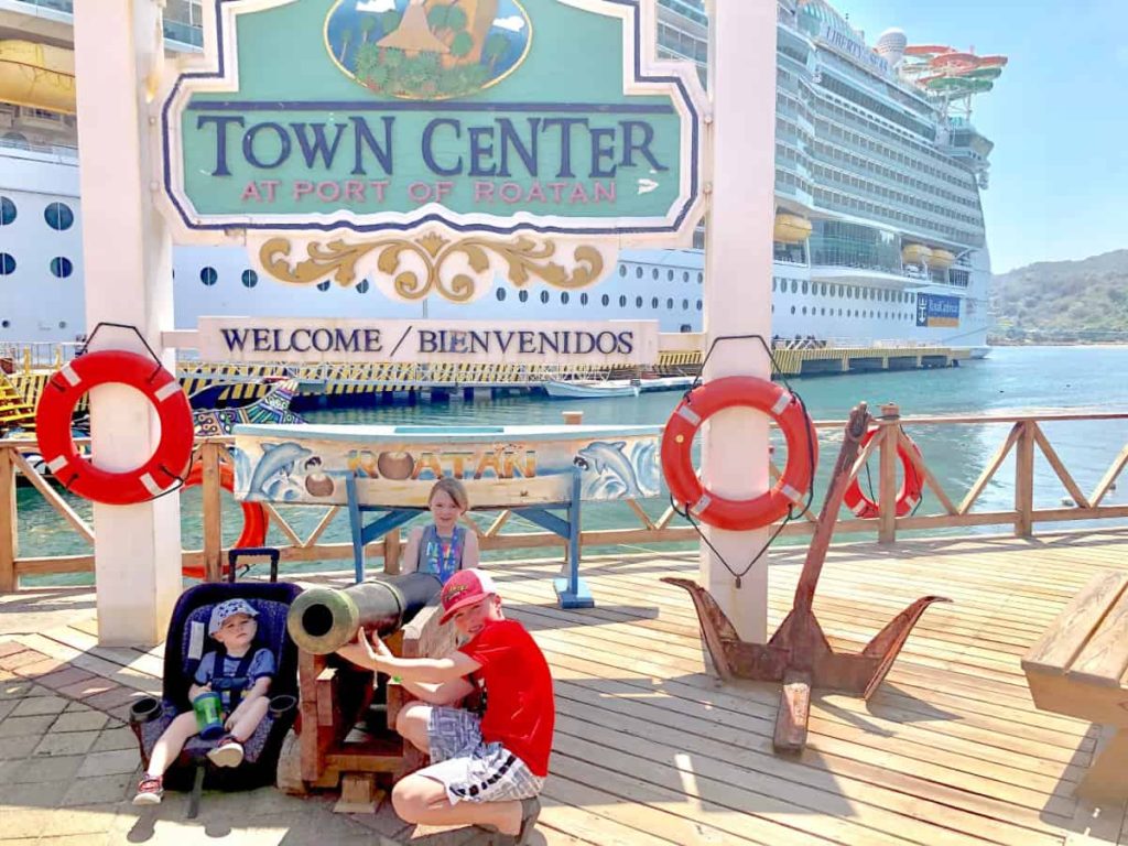 3 kids standing in front of the town center port of roatan with a cruise ship in the background. 