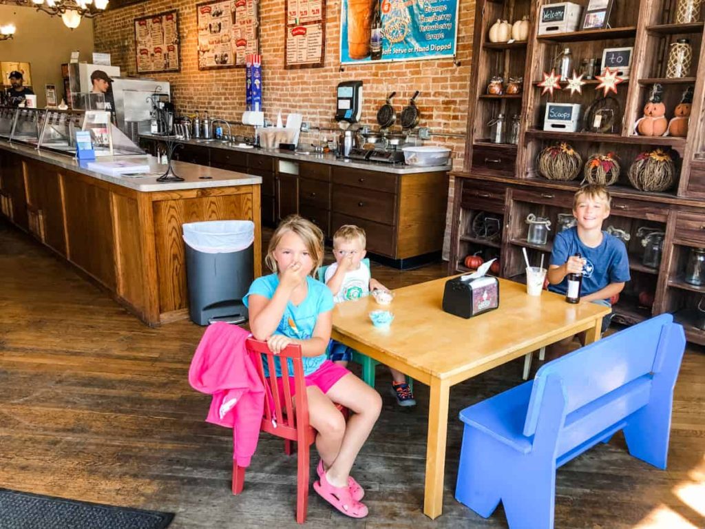 3 kids sitting at a small table made for kids inside of an ice cream shop. 