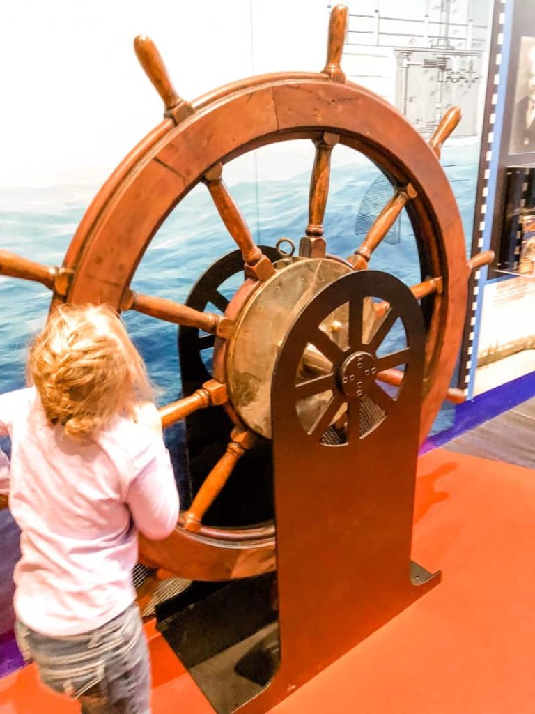 Girl working hard to turn a giant wooden ship wheel.