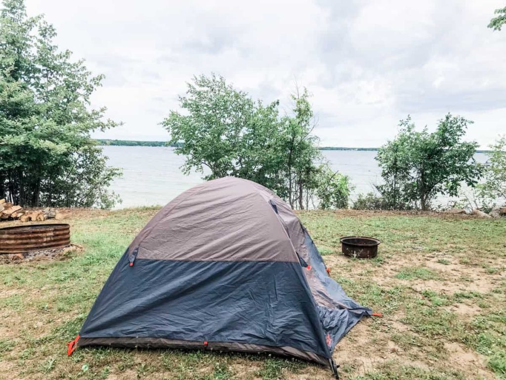 small 3 person tent set up at a site that is right next to a lake. 