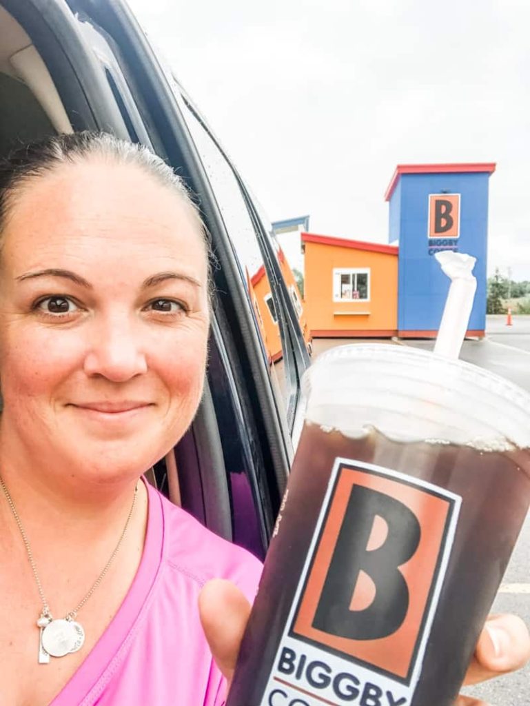 Women taking selfie of Biggby Cold Coffee Cup with straw with a tiny shed like Biggby Coffee Drive through in the background.