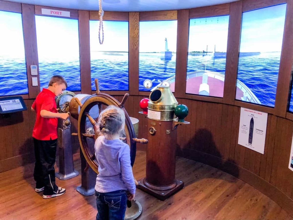 Boy and Girl playing with a virtual ship. Digital screens look like the lake, and real boat parts are in place to make them feel like they are driving a real ship.
