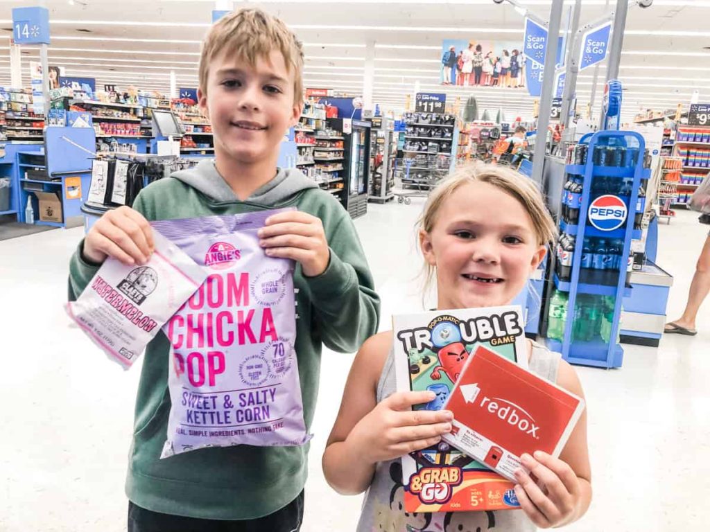 Boy holding Boom Chicka Pop and old fashion hard candy, and girl holding Travel Trouble game and Red Box DVD. Standing in front of a store checkout. 