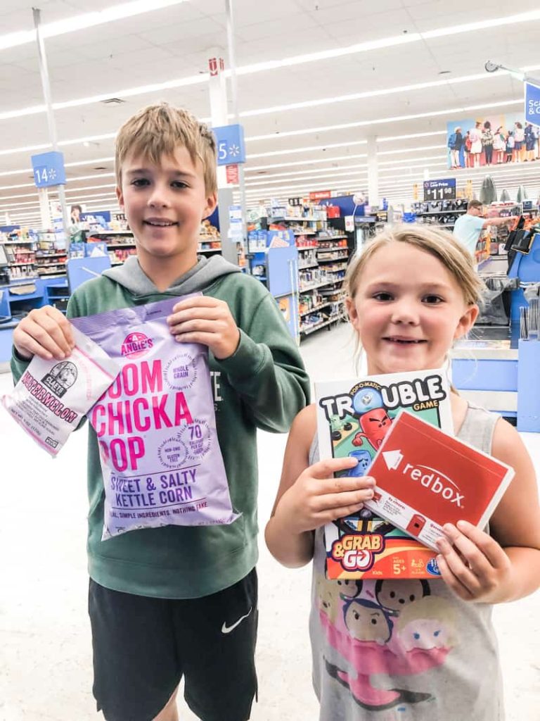 Boy and girls standing in front of a store check out area with a bag of boom chicka pop old fashion hard candy, travel trouble and red box dvd
