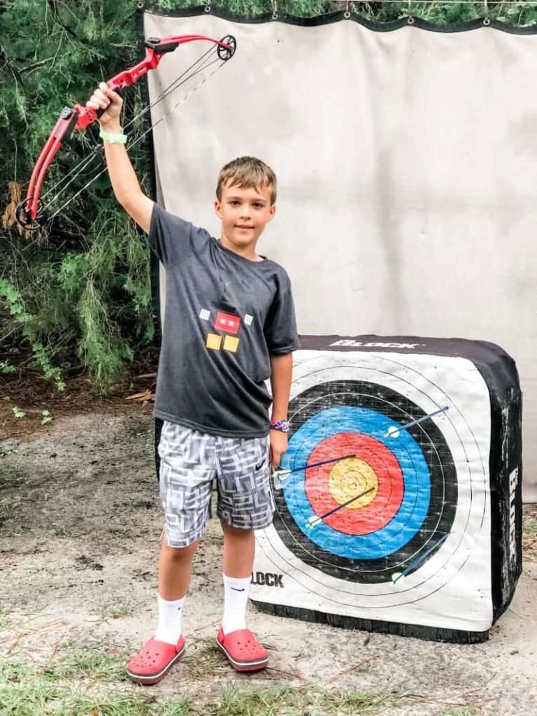 Boy standing in front of target that has 4 arrows in it. Victoriously holding the bow above his head. 