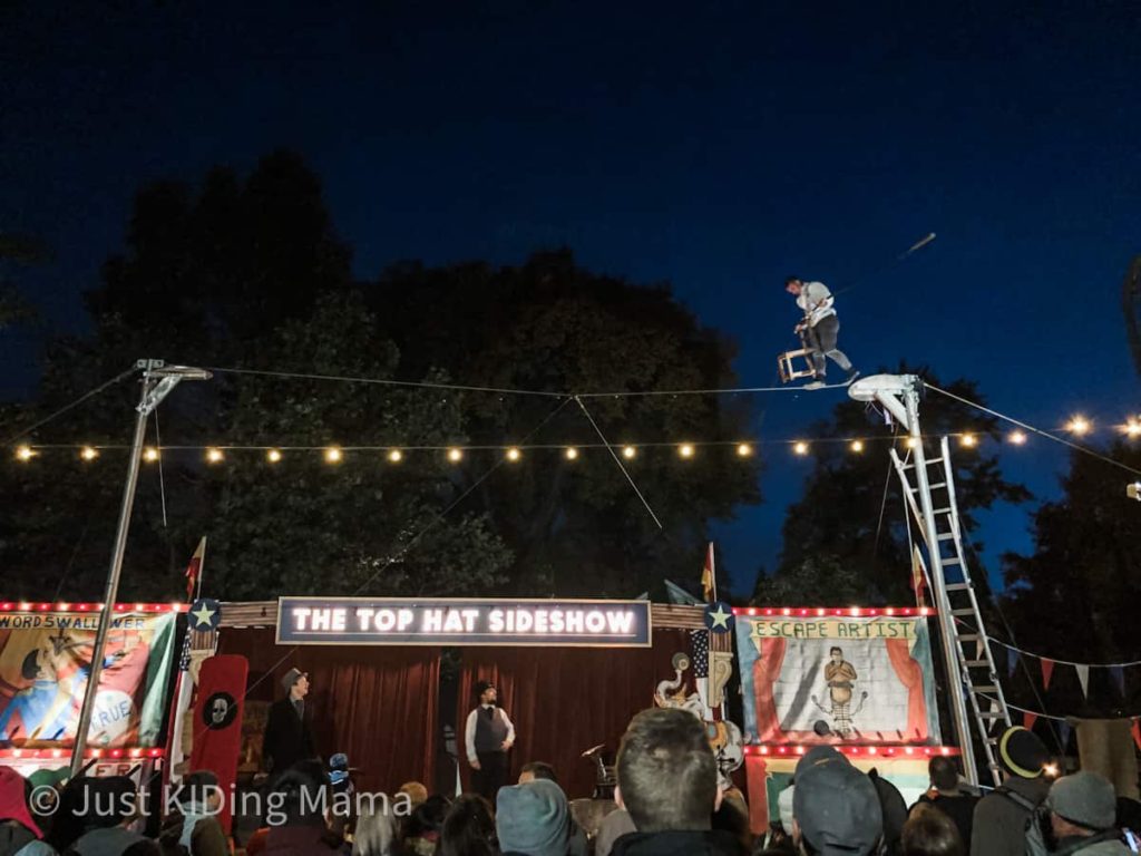 Crowd gathers at a circus themed side show while watching a man on a tightrope. 