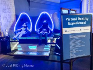 2 kids sitting in chair pods with VR headsets on. The chair pods have glowing blue lights around the outside. 