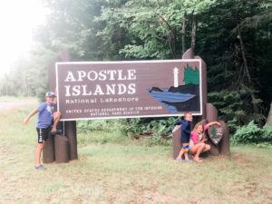 2 boys and a girl starnding in front of the Apostle Island National Lakeshore brown sign with lighthouse and lake painted on it.