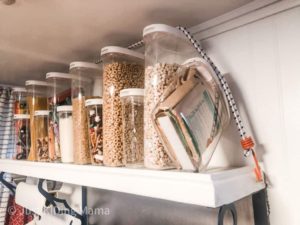 Clear food storage containers with white tops all filled with cereal, nuts, oatmeal and marshmallows on a shelf hook and look attached as well as a bungee cord