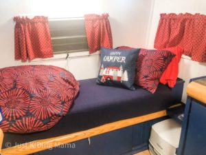 Back bed of a camper. Navy blue cushion, which wall, red curtains, and Happy Camper pillow