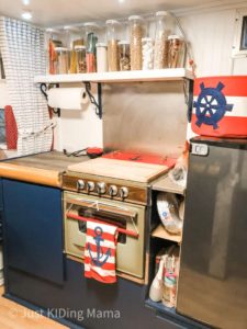 Vintage Camper Interior. Green oven, blue lower cabinets, and a row of food in storage containers above the oven. 