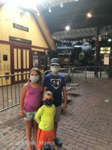 2 boys and a girl wearing medical masks, standing inside a train museum in front of real train cars.