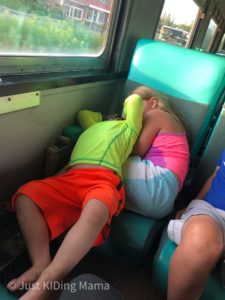 toddler boy throwing himself backwards ontop of his sister who is looking embarrassed while sitting on train car seat