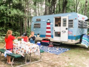 2 boys and a girl are eating a picnic dinner. Behind them is a vintage camper painted blue with a lighthouse on it.