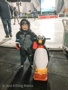 Boy in snow gear, skates, and hockey helmet standing next to a plastic penguin with handle bars with an outdoor ice rink behind them