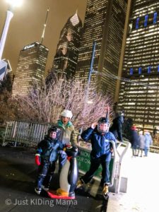 2 boys and a girl dressed in snow gear, skates, and hockey helmets standing next to an ice rink with the Chicago skyline in the background