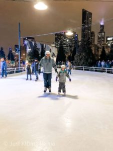 Dad and girl, both wearing hockey helmets ice skating on an outdoor rink with the Chicago Skyline behind them. 